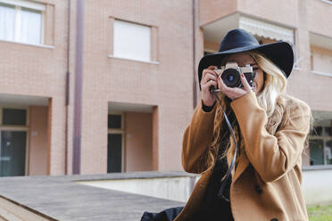 Junge Frau mit Hut filmt durch die Kamera gegen ein Gebäude - FMOF01356