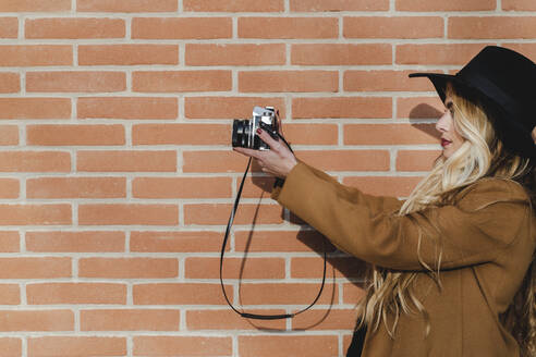 Junge Frau mit blonden Haaren hält die Kamera gegen eine Backsteinmauer an einem sonnigen Tag - FMOF01361