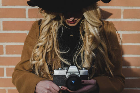 Junge blonde Frau mit Kamera an einer Mauer stehend - FMOF01364