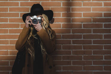 Frau mit Hut, die durch die Kamera filmt, während sie gegen eine Backsteinmauer steht - FMOF01365