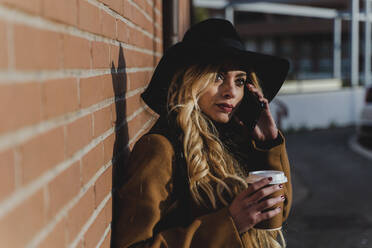 Blond woman talking on smart phone while holding coffee cup during sunny day - FMOF01366