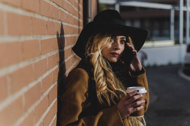 Blonde Frau spricht mit ihrem Smartphone und hält eine Kaffeetasse an einem sonnigen Tag - FMOF01366