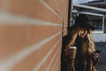 Blond woman with coffee cup using phone while standing by brick wall - FMOF01367