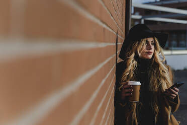 Young woman in hat with coffee cup standing by brick wall while looking away - FMOF01368
