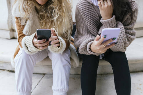 Freundinnen in warmer Kleidung, die auf einer Treppe sitzend ein Mobiltelefon benutzen - JRVF00206