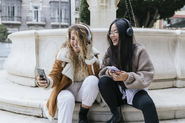 Lachende weibliche Freunde, die ein Selfie mit Kopfhörern machen, während sie gegen einen Springbrunnen sitzen - JRVF00207