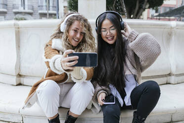 Smiling female friends taking selfie wearing headphones while sitting against fountain - JRVF00208