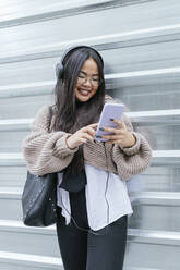 Junge Frau hört Musik über ihr Smartphone, während sie an einer Metallwand steht - JRVF00211