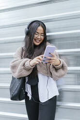 Young woman listening music through smart phone while standing against metal wall - JRVF00211