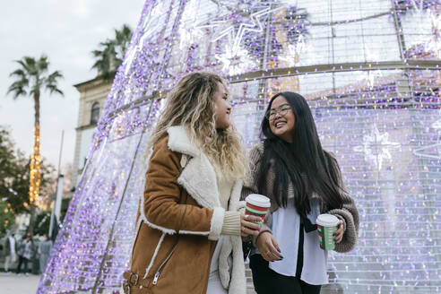 Cheerful female friends holding coffee cup while walking by Christmas decoration at street - JRVF00223