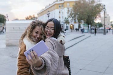 Cheerful female friends taking selfie through smart phone while embracing on sidewalk in city - JRVF00225