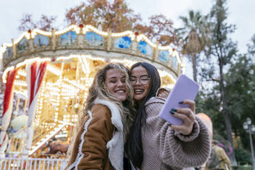 Multi-ethnic female friends talking selfie through mobile phone against illuminated carousel - JRVF00226