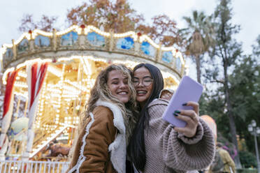 Multi-ethnische Freundinnen sprechen Selfie durch Handy gegen beleuchtetes Karussell - JRVF00226