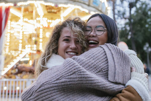 Lachende Freundinnen, die sich vor einem beleuchteten Karussell umarmen - JRVF00227