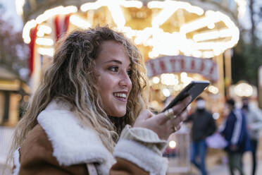 Junge Frau, die eine Sprachnachricht über ihr Smartphone sendet, während sie vor einem beleuchteten Karussell steht - JRVF00228
