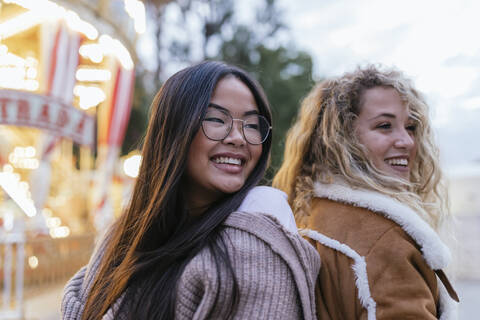 Multi-ethnische Freundinnen schauen weg, während sie Rücken an Rücken vor einem beleuchteten Karussell stehen, lizenzfreies Stockfoto