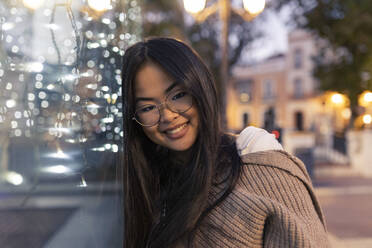 Glückliche junge Frau, die sich in der Abenddämmerung an eine beleuchtete Glaswand lehnt - JRVF00232
