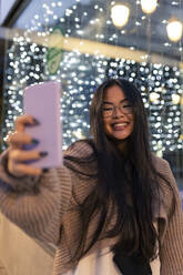 Fröhliche junge Frau, die ein Selfie mit ihrem Smartphone vor einer beleuchteten Glaswand macht - JRVF00236