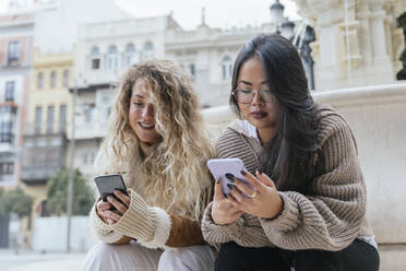 Multiethnische Freundinnen benutzen ihr Smartphone, während sie am Brunnen sitzen - JRVF00239