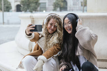 Cheerful female friends taking selfie through smart phone while listening music over headphones - JRVF00240