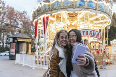Multi-ethnische Freundinnen machen ein Selfie vor einem beleuchteten Karussell im Vergnügungspark - JRVF00245