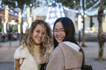 Junge Freundinnen lächelnd vor beleuchteter Weihnachtsdekoration in der Abenddämmerung - JRVF00246