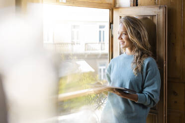 Lächelnde Frau mit digitalem Tablet, die zu Hause aus dem Fenster schaut - SBOF02625