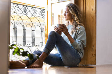 Frau hält Kaffeetasse und schaut durch das Fenster zu Hause - SBOF02630