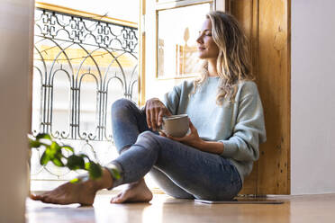 Woman with eyes closed holding coffee cup while sitting on floor at home - SBOF02632