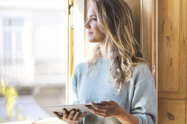 Smiling thoughtful woman with digital tablet looking out of window at home - SBOF02635