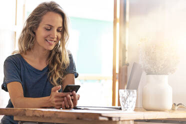 Lächelnde Freiberuflerin, die im Büro zu Hause ein Mobiltelefon benutzt - SBOF02654