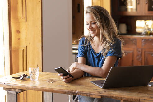 Lächelnde Geschäftsfrau mit Smartphone am Tisch im Wohnzimmer - SBOF02659