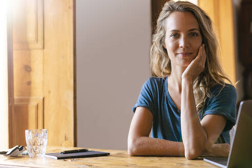 Lächelnde Geschäftsfrau mit Hand am Kinn neben dem Laptop im Heimbüro - SBOF02664