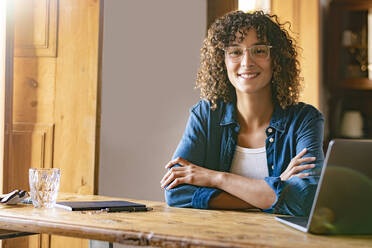 Happy young businesswoman with arms crossed by laptop at home office - SBOF02665