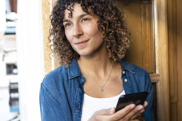 Curly woman with smart phone looking away at home - SBOF02668