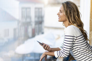 Woman with smart phone leaning on handrail in balcony - SBOF02674