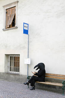 Eine Frau im Krähenkostüm benutzt ein Mobiltelefon, während sie auf der Bank einer Bushaltestelle sitzt - PSTF00779
