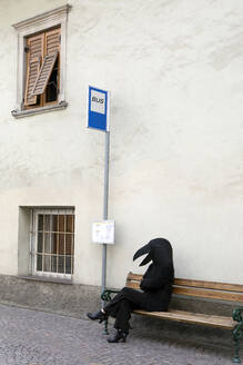 Frau im Vogelkostüm sitzt auf der Bank einer Bushaltestelle - PSTF00781