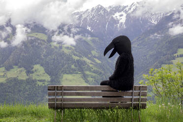 Frau im Krähenkostüm sitzt auf einer Bank vor einer Bergkette - PSTF00793