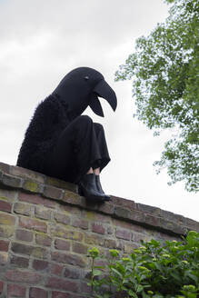 Frau im Krähenkostüm sitzt auf Stützmauer gegen Himmel - PSTF00796