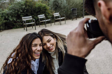 Young man photographing cheerful female friends at park - XLGF01112