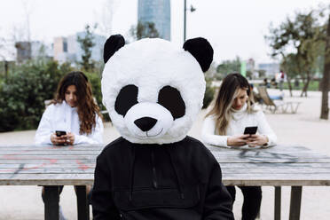 Young man with panda mask sitting against female friends at park - XLGF01118