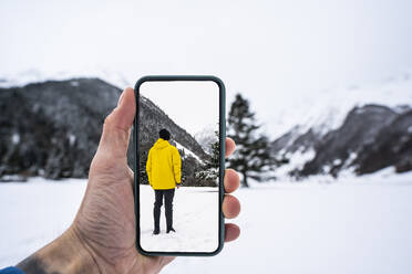 Fotografierende Männerhand, während ein männlicher Freund in einem verschneiten Tal steht - DAMF00679