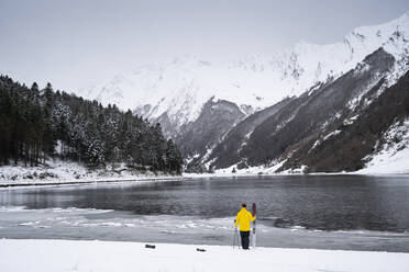 Mann mit Ski am schneebedeckten Seeufer vor den Bergen - DAMF00684