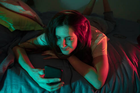 Smiling woman using mobile phone while lying on front in bedroom - AFVF08150