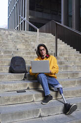 Young man with leg prosthesis using laptop while sitting on steps - VEGF03735