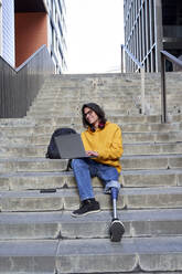 Smiling young disabled man with artificial limb using laptop while sitting on steps in city - VEGF03736