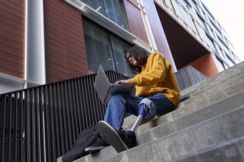 Young man with artificial limb using laptop while sitting on steps against building - VEGF03738