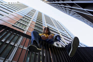 Behinderter junger Mann mit Beinprothese sitzt auf einem Metallgeländer vor einem Hochhaus - VEGF03740