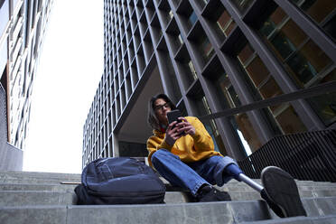 Junger Mann mit Beinprothese benutzt Mobiltelefon, während er auf den Stufen eines Hochhauses sitzt - VEGF03742